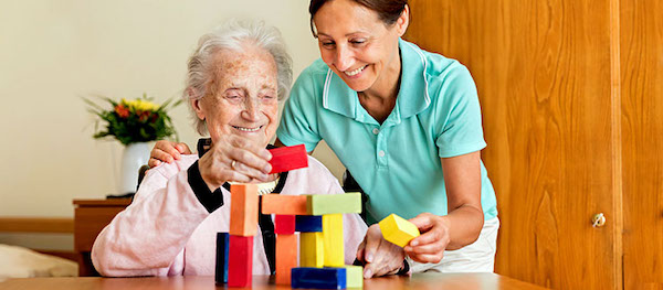 An elderly female playing with building blocks with the assistance of a healthcare worker